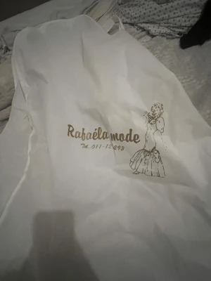 Vit långklänning från Rafaëla Mode - Elegant vit bröllops klänning  med vackra blommiga spetsdetaljer och pärlor. Klänningen har omlottband i midjan och är ärmlös med en snygg v-ringad överdel. De finns bara 3 sånna här klänningar för den har varit i en runway som modellklänning 