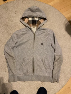 Grå hoodie från Burberry M - Snygg grå zip-hoodie från Burberry Brit i storlek M. Klassisk rutmönstrad insida på huvan, broderad Burberry-logga på bröstet och två stora fickor fram. Tillverkad i mjuk bomullsblandning, perfekt för chill dagar.