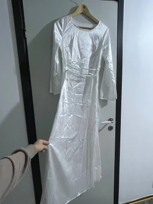 Vit långärmad satin klänning - Säljer en elegant vit långklänning med långärmade och rund halsringning. Klänningen har ett smalt satinband i midjan som ger en snygg siluett. Materialet är glansigt och känns lyxigt, perfekt för dig som vill sticka ut med en stilren look.