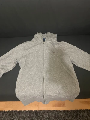 Grå zip-hoodie från Gant - Snygg grå hoodie från Gant med dragkedja hela vägen, huva med dragsko och två stora fickor framtill. Liten broderad Gant-logga på bröstet. Mjuk bomullskänsla och klassisk passform, perfekt för chill dagar.