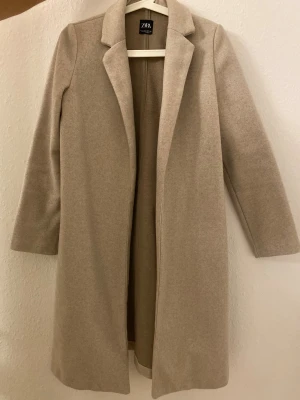 Beige kappa från Zara i storlek S - Snygg beige kappa från Zara med klassisk krage och långärmad design. Jackan har en rak och stilren form utan synliga knappar eller dragkedja, vilket ger en clean look. Perfekt för dig som gillar minimalistisk stil och neutrala färger.