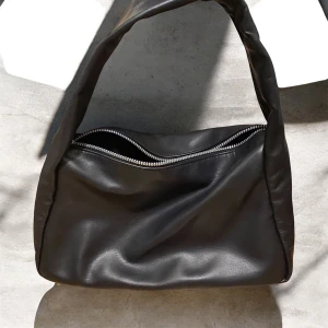 Svart axelväska i skinnlook - Stilren svart axelväska med mjuk, slät yta i skinnimitation. Väskan har en bred axelrem och en silvrig dragkedja upptill. Perfekt för dig som gillar minimalistisk och trendig design.