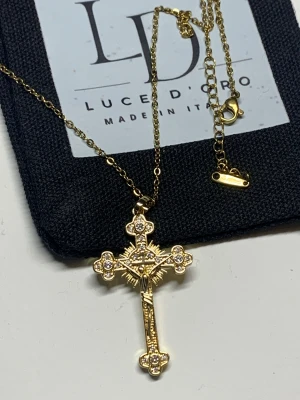 Guldfärgat korshalsband Luce D'Oro - Snyggt guldfärgat halsband från Luce D'Oro med ett detaljerat korshänge. Korset är dekorerat med gnistrande stenar och har en klassisk kedja i rostfritt stål. Perfekt för dig som gillar statement-smycken med religiös touch.