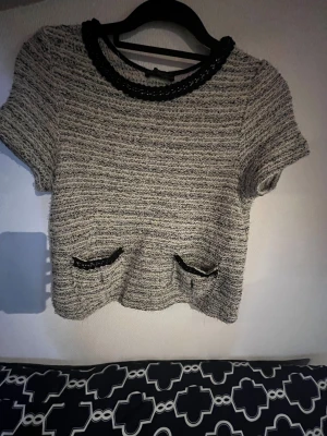 Grå stickad topp med kedjedetaljer - Säljer en grå stickad topp med korta ärmar och två små fickor framtill, dekorerade med svarta kedjedetaljer. Toppen har en rund halsringning med svart kedja som extra detalj. Perfekt för dig som gillar en chic och trendig look.