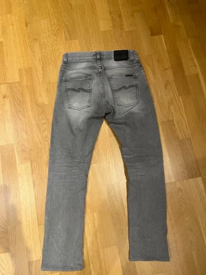 Grå jeans från Nudie, straight fit - Snygga grå jeans från Nudie med klassisk femficksdesign och diskret tvättad look. Modellen har raka ben och normal passform, med svarta detaljer på bakfickorna. Perfekt för dig som gillar stilrena och tidlösa jeans.