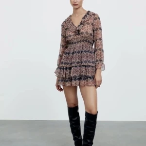 Mönstrad kortklänning med volanger - Supersöt kortklänning med blommigt mönster i bruna, röda och blå toner. Klänningen har volanger, v-ringning med rysch och långa ärmar i transparent material. Perfekt för dig som gillar boho-vibe och vill sticka ut med coola detaljer. Jätte snygg nu till julen/vinter då de mörkare färgerna