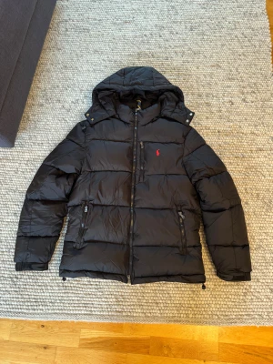 Polo Ralph Lauren jacka - Svart pufferjacka från Polo Ralph Lauren med huva, dragkedja och två fickor framtill. Jackan har det klassiska röda Polo-loggan broderad på bröstet och är perfekt för kalla dagar. Fylld med dun och har justerbara detaljer vid ärmslut och nederkant.