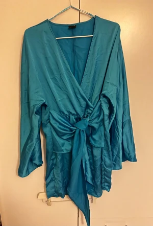 Turkos klänning med knytband - Säljer en turkos klänning i glansigt satinliknande material. Klänningen har långa, vida ärmar och ett brett knytband i midjan som ger en snygg siluett. Perfekt för dig som vill sticka ut med färg och cool form.