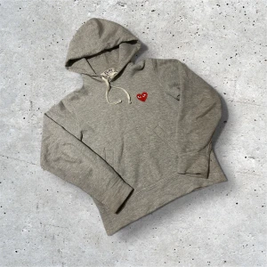 Grå hoodie Comme des Garçons Play - Snygg grå hoodie från Comme des Garçons Play med klassiskt rött hjärta med ögon broderat på bröstet. Mjuk bomullsmix, dragsko i huvan och känguruficka framtill. Perfekt för en chill och stilren look.