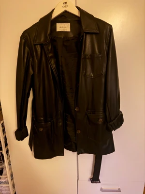 Svart skinnjacka från Stradivarius - Snygg svart skinnjacka från Stradivarius med klassisk krage, knappar framtill och tre fickor. Jackan har bälte i midjan och långa ärmar. Perfekt för dig som vill ha en stilren och edgy look.
