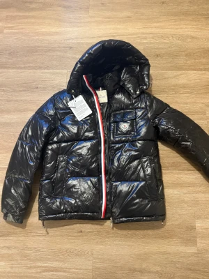 Svart Moncler pufferjacka med huva - Svart pufferjacka från Moncler med glansig finish, huva och ikonisk logga på bröstet. Jackan har en röd, vit och blå dragkedja, praktiska fickor och quiltad design. Perfekt för dig som vill ha en snygg och varm jacka med street-vibe.