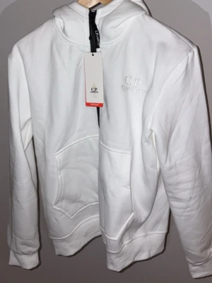 Vit hoodie från C.P. Company - Vit hoodie från C.P. Company med broderad logga på bröstet och klassiska goggles på huvan. Mjuk bomullskvalitet, känguruficka framtill och dragkedja vid halsen. Perfekt för en streetwear-look.