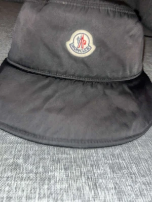 Svart bucket hat från Moncler - Stilren svart bucket hat från Moncler med klassisk logotyp framtill. Hatten är tillverkad i ett glansigt, vattenavvisande material och har en bred skärm som skyddar mot solen. Perfekt accessoar för att lyfta din outfit. ÄKTA!!!