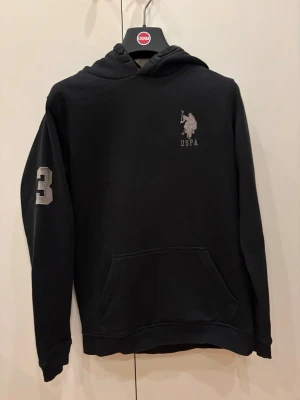 Svart hoodie från US Polo Assn - Svart hoodie med USPA-logga på bröstet och en stor ficka framtill. Siffran 3 är broderad på ena ärmen. Klassisk huva och ribbade muddar. Perfekt för en chill och sportig stil.