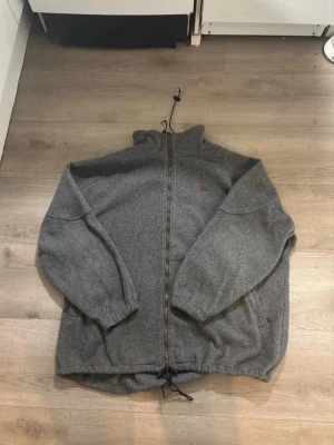 Grå stickad fleecejacka Fjällräven - Mysig grå stickad fleecejacka från Fjällräven med hel dragkedja och hög krage. Jackan har dragsko nertill och broderad logga på bröstet. Perfekt för kyliga dagar och enkel att matcha med andra plagg.