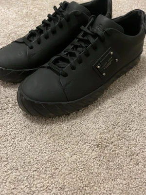 Svarta sneakers från Philipp Plein - Stilrena helsvarta sneakers från Philipp Plein med mattsvart finish och metallplatta på sidan. Skorna har snörning, rund tå och platt sula. Perfekta för dig som gillar en clean och modern look.