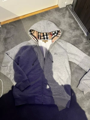 Äkta zip hoodie från burberry  - Snygg grå hoodie från Burberry med dragkedja och klassiskt rutigt foder i huvan. Mjuk bomullsmix, ribbade muddar och fickor framtill. Perfekt för dig som vill ha en stilren och exklusiv look med en twist av det ikoniska Burberry-mönstret.