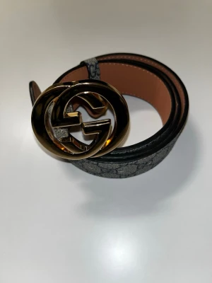 GG bälte - Snyggt bälte från GG med klassiskt mönster och stort runt guldspänne. Bältet är svart och brunt med insida i ljusbrunt skinn. Perfekt accessoar för att lyfta din outfit.