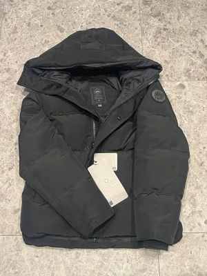 Svart pufferjacka från Canada Goose - Säljer en svart pufferjacka från Canada Goose med huva och diskret logotyp-patch på ärmen. Jackan har dragkedja och tryckknappar framtill samt två sidofickor. Perfekt för kalla vinterdagar och riktigt snygg till streetwear-looken.