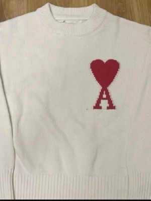 Vit stickad tröja från Ami - Vit stickad tröja från Ami med röd hjärtlogga och bokstaven A på bröstet. Tröjan har rund halsringning och ribbade muddar vid ärmslut och nederkant. Perfekt för en stilren och trendig look.