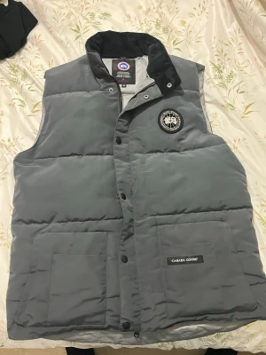 Grå dunväst från Canada Goose M - Säljer en grå dunväst från Canada Goose med svart krage och tydlig logga på bröstet. Västen har två stora fickor framtill och stängs med både dragkedja och tryckknappar. Perfekt för lager-på-lager och riktigt snygg till höst- och vinterlooken.