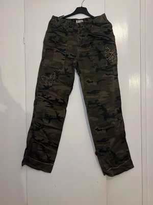 Camo cargobyxor med strassdetaljer - Snygga cargobyxor i camouflage-mönster från Cecil. Byxorna har flera fickor och coola strassdetaljer på fickorna framtill och baktill. Vida ben och klassisk cargostil, perfekta för en streetwear-look. Materialet är bomull för skön känsla.