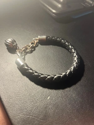 Svart flätat läderarmband med berlock - Snyggt svart armband i flätat läder med silverfärgade metalldetaljer och en rund berlock. Armbandet har en enkel kedja och stängning i metall. Perfekt accessoar för en stilren look.