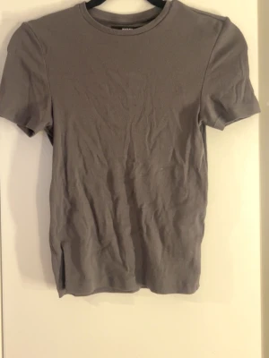 Grå ribbad t-shirt - En enkel grå t-shirt med ribbad struktur och rund hals. T-shirten har korta ärmar och en klassisk passform som funkar till det mesta. Perfekt basplagg för garderoben.
