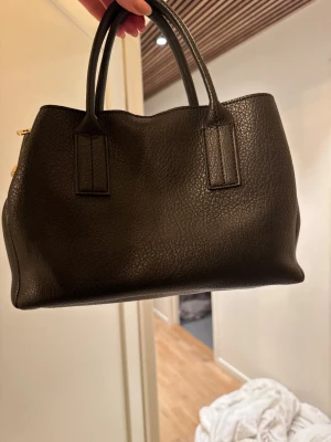 Svart handväska i skinn med guldiga detaljer - Snygg svart handväska i äkta skinn med två handtag och guldiga metalldetaljer. Väskan har en stilren, rektangulär form och en klassisk look som passar till många outfits. Perfekt för dig som gillar minimalistisk och tidlös design.