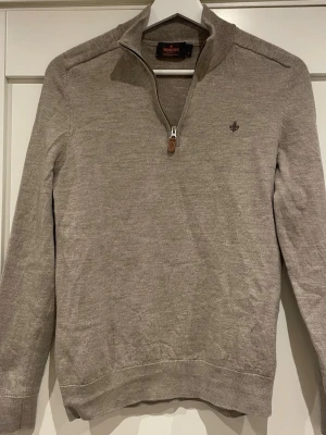 Beige stickad tröja från Morris - Snygg beige stickad tröja från Morris i 100% merinoull. Tröjan har halv dragkedja med läderdetalj, ribbad krage och muddar samt Morris logga broderad på bröstet. Perfekt för dig som gillar klassisk och stilren design.