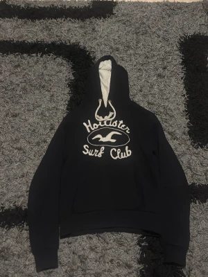 Svart Hollister hoodie  - Svart hoodie från Hollister med vit broderad text 'Hollister Surf Club' och vågmotiv på bröstet. Klassisk passform och mjukt material som känns skönt mot huden. St:S men sitter mer som en xs. Priser är så klart inte hugget i sten
