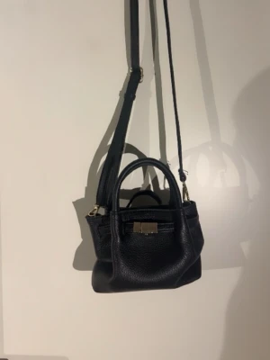Svart miniaxelväska med gulddetaljer - Snygg svart axelväska i miniformat med justerbar axelrem och guldfärgat spänne framtill. Väskan har både handtag och axelrem, vilket gör den enkel att bära på olika sätt. Tillverkad i strukturerat konstläder för en trendig look.