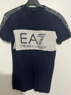 EA7 Emporio Armani blå t-shirt - Snygg marinblå t-shirt från EA7 Emporio Armani med vit panel och stor logga framtill. T-shirten har korta ärmar och logotypband längs axlarna. Tillverkad i mjuk bomull för en bekväm känsla och modern look. Storlek s passar xs