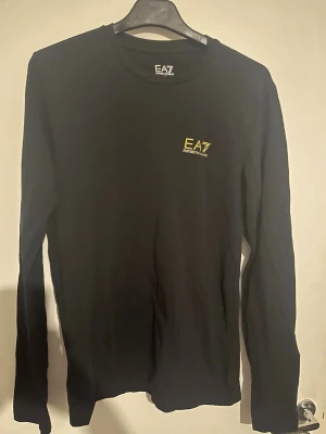 Svart långärmad topp från EA7 - Svart långärmad topp från EA7 Emporio Armani med rund halsringning och liten gul logga på bröstet. Tillverkad i mjuk bomull som känns skön mot huden. Perfekt för en stilren och sportig look. Storlek xs (170) barnstorlek 