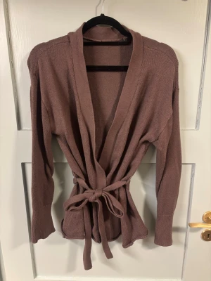 Mauve stickad omlottkofta med knytskärp - Säljer en stickad omlottkofta i en snygg brun nyans. Koftan har långa ärmar, djup V-ringning och ett matchande knytskärp i midjan för en schysst siluett. Perfekt att slänga på sig över en topp eller t-shirt.