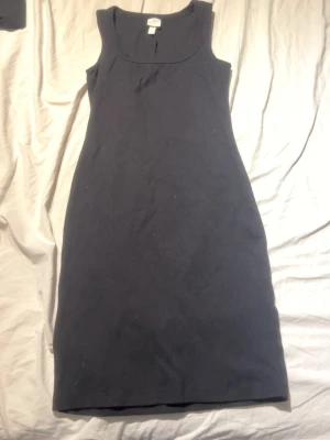 Svart ärmlös klänning i bomull - En enkel och stilren svart klänning i bomull med ärmlös design och rundad halsringning. Klänningen har en rak passform och slutar strax ovanför knäna, perfekt för en clean och klassisk look.