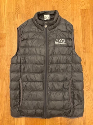 Svart dunväst från Emporio Armani - Snygg grå dunväst från Emporio Armani EA7 med quiltad design och dragkedja framtill. Västen har hög krage, två sidofickor och logga på bröstet. Perfekt för lager-på-lager och streetwear-stil. Vafan må se lite skrynklig ut men det är pga att den legat länge i garderob, skrynklorna försvinner naturligt snabbt vid användning. 