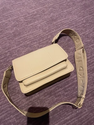 Beige axelväska från NA-KD - Snygg beige axelväska från NA-KD med bred axelrem där loggan är präglad. Väskan har en stilren, rektangulär form och är gjord i konstläder. Perfekt för dig som vill ha en trendig och praktisk väska med plats för det viktigaste.