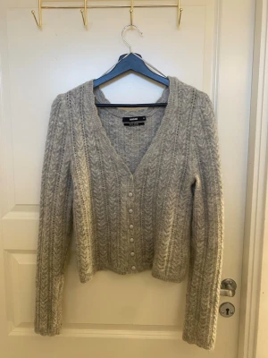 Grå kabelstickad kofta från Bik Bok - Mysig grå kabelstickad kofta från Bik Bok med v-ringning och pärlemorknappar framtill. Koftan har lång ärm och ett klassiskt stickat mönster som ger en trendig look. Perfekt att slänga över en topp för extra värme.