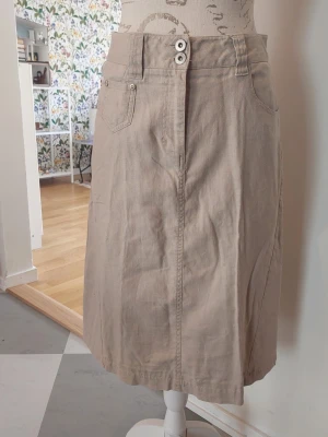 Beige långkjol med fickor och knappar - Stilren beige långkjol med två knappar i midjan och praktiska fickor fram. Kjolen har en rak passform och är tillverkad i ett mjukt bomullsmaterial. 