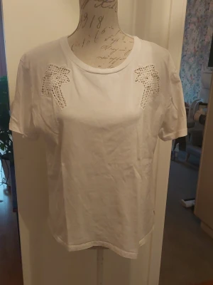 Vit t-shirt med spetsdetaljer - Vit t-shirt i bomull med korta ärmar och rund halsringning. Framtill finns dekorativa spetsdetaljer i form av palmblad vid axlarna. Perfekt för en avslappnad och stilren look.