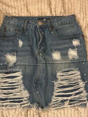 Blå jeanskjol från Boohoo - Trendig blå jeanskjol från Boohoo med kraftiga slitningar och fransad nederkant. Kjolen har pärldetaljer, klassisk femficksdesign och hög midja. Perfekt för dig som gillar en edgy och avslappnad stil.