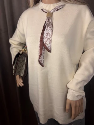 Brun paisleymönstrad halsduk - Snygg halsduk i sidenliknande material med paisleymönster i brunt och vitt. Halsduken har en smal, avlång form och är kantad med brun ram. Den hålls ihop med ett guldfärgat metallspänne för en trendig touch.