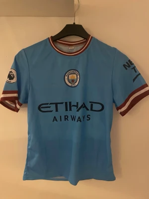 Manchester City matchtröja De Bruyne - Snygg ljusblå Manchester City matchtröja med De Bruyne och nummer 17 på ryggen. Tröjan har klubbmärke, Etihad Airways-tryck och Premier League-logga på ärmen. Detaljer i vinrött och vitt på ärmslut och krage. Perfekt för fotbollsfans!