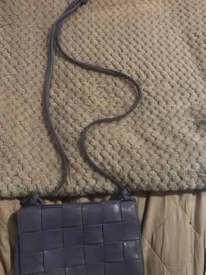 Lila flätad axelväska  - Trendig lila axelväska med flätad framsida. Väskan är gjord i skinnimitation och har en stilren, rektangulär form. Perfekt för dig som vill ha en snygg accessoar som sticker ut. Perfekt att lägga nycklar, mobil, plånbok etc i. 