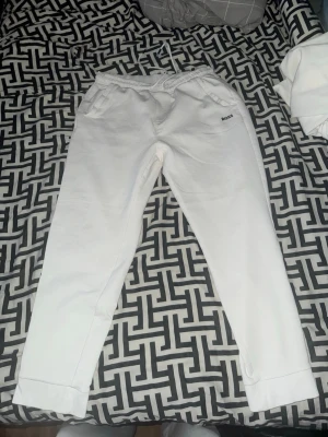 Vita mjukisbyxor från BOSS - Säljer ett par vita mjukisbyxor från BOSS med elastisk midja och dragsko. Byxorna har fickor fram och en bakficka med BOSS-logga. Sköna muddar vid bensluten och stilren design, perfekta för chill dagar. Använd enstaka gånger.