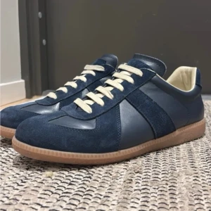 Mörkblå Maison Margiela sneakers med mockadetaljer - Snygga mörkblå sneakers med ovandel i både mocka och läder, beige innersida och kontrasterande vita skosnören. Yttersulan är i gummi med naturfärg. Perfekta för dig som gillar stilrena och klassiska sneakers med en twist.