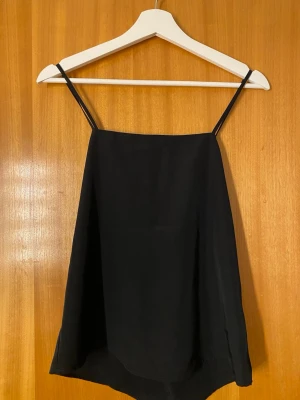 Svart linnetopp med smala axelband -  svart linnetopp med smala axelband och rak passform. Toppen har en enkel design framtill, banden är korsade i ryggen men går även att ha de korsade i fram. Haft på mig den vid ett tillfälle. För liten!