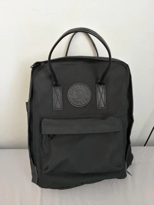 Svart Kånken ryggsäck från Fjällräven - Klassisk svart Kånken ryggsäck från Fjällräven med rund logotyp framtill. Ryggsäcken har ett stort huvudfack, en framficka med dragkedja och två handtag upptill. Tillverkad i slitstarkt syntetmaterial och har justerbara axelband. Helt ny endast testad. Köpt för 1900kr