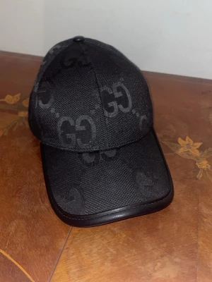 Svart GG-keps från Gucci - Snygg svart keps från Gucci med GG-monogram i ton-i-ton över hela kepsen. Klassisk böjd skärm och detaljer i svart läder runt kanten. Tillverkad i ett slitstarkt tyg som ger en exklusiv känsla och stil.
